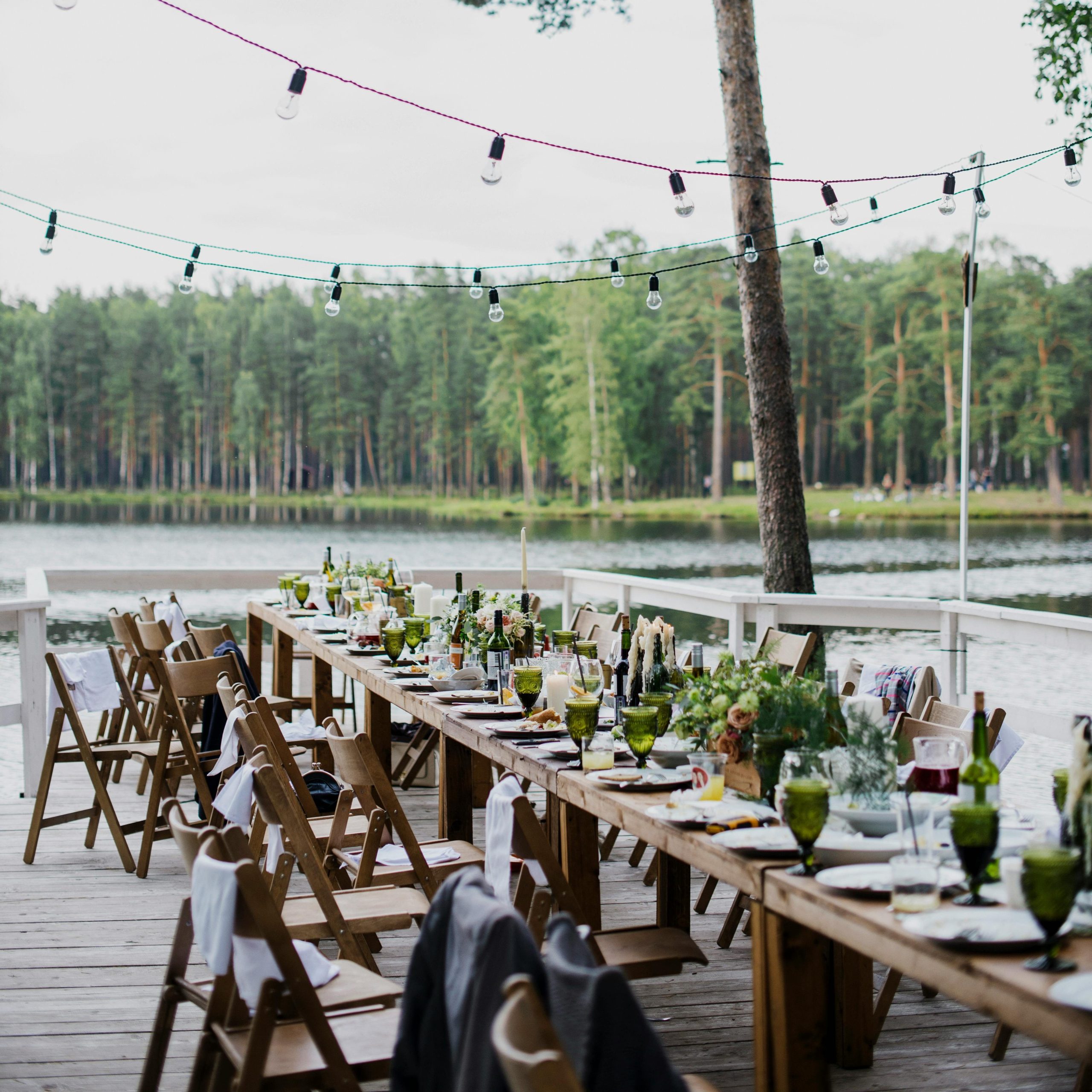 Wooden terrace with green trees and tables located near lake preparing for festive romantic party in nature under blue sky
